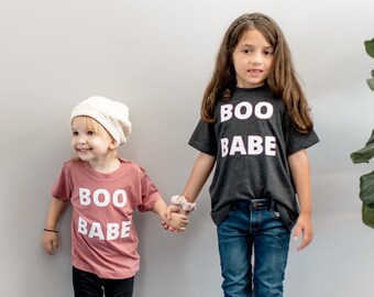 Könnte beinhalten: Ein altrosa T-Shirt mit weißem Schriftzug BOO BABE, schwarze Leggings und Leoparden-Stiefel. Ein anthrazitfarbenes T-Shirt mit weißem Schriftzug BOO BABE, blaue Jeans und beige Wildleder-Stiefel mit cremefarbenem Fleecefutter.