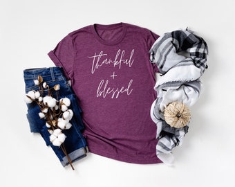 Puede incluir: Una camiseta de manga corta en color ciruela jaspeado con el texto cursivo blanco "thankful + blessed" impreso en el frente. La camiseta se muestra junto a unos vaqueros azul oscuro, una rama de algodón blanco, una bufanda de cuadros en blanco y negro y una pequeña calabaza de cuerda.