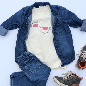 K&ouml;nnte beinhalten: Ein cremefarbenes T-Shirt mit dem Text "HE HAS RISEN JUST AS HE SAID" in Schwarz und einer rosa Sonnengrafik. Das Shirt wird mit einer Jeansjacke, einer blauen Jeans und grauen und braunen Turnschuhen kombiniert. Ein l&auml;ssiges Outfit.