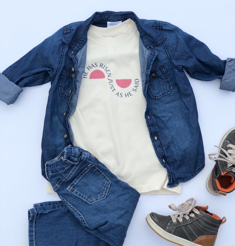 K&ouml;nnte beinhalten: Ein cremefarbenes T-Shirt mit dem Text "HE HAS RISEN JUST AS HE SAID" in Schwarz und einer rosa Sonnengrafik. Das Shirt wird mit einer Jeansjacke, einer blauen Jeans und grauen und braunen Turnschuhen kombiniert. Ein l&auml;ssiges Outfit.