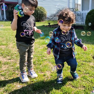 May include: Two young children playing outdoors on a sunny day, surrounded by bubbles. One child wears a black t-shirt with a bunny graphic, olive green pants, and sneakers. The other child wears a plaid shirt, a similar bunny graphic t-shirt, and blue jeans.