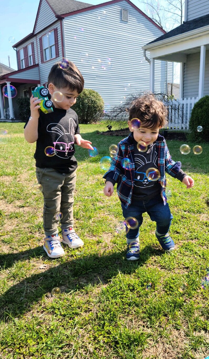May include: Two young children playing outdoors on a sunny day, surrounded by bubbles. One child wears a black t-shirt with a bunny graphic, olive green pants, and sneakers. The other child wears a plaid shirt, a similar bunny graphic t-shirt, and blue jeans.