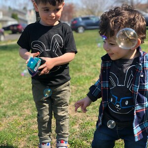 May include: Two young children playing outdoors on a sunny day. One child wears a black t-shirt with a white bunny graphic and khaki pants. The other child wears a similar shirt with glasses and a blue plaid shirt. Bubbles float in the air.