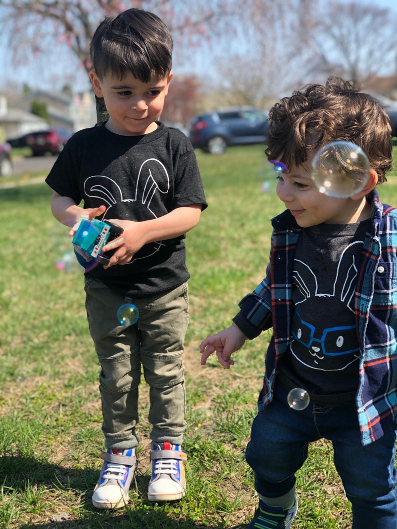 May include: Two young children playing outdoors on a sunny day. One child wears a black t-shirt with a white bunny graphic and khaki pants. The other child wears a similar shirt with glasses and a blue plaid shirt. Bubbles float in the air.