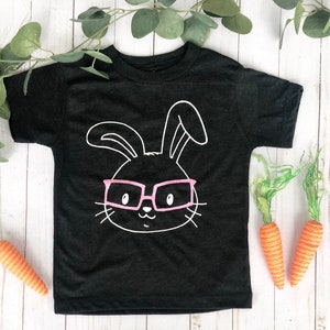 May include: Black toddler t-shirt featuring a white outline of a bunny face wearing pink glasses. Two orange carrots and green foliage are placed near the shirt on a white wooden surface. A fun Easter-themed design.