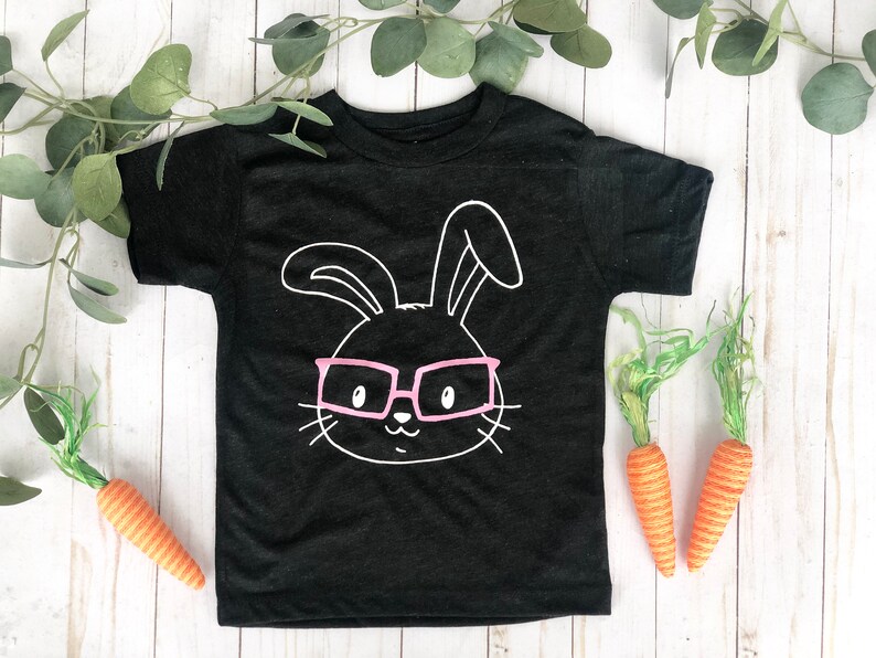 May include: Black toddler t-shirt featuring a white outline of a bunny face wearing pink glasses. Two orange carrots and green foliage are placed near the shirt on a white wooden surface. A fun Easter-themed design.
