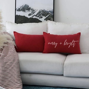 May include: Two rectangular throw pillows in a deep red fabric. One pillow is plain, while the other features the white cursive text merry + bright centered on the front. The pillows are positioned on a white sofa, providing a clear view of the red color and the white lettering on the decorative accent piece.