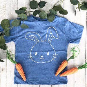 May include: A blue t-shirt with a white bunny face design. The shirt is decorated with two orange carrots and green foliage. The shirt is on a white wooden surface. The design is simple and suitable for Easter.