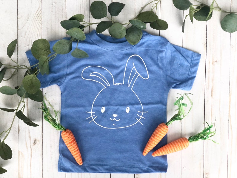 May include: A blue t-shirt with a white bunny face design. The shirt is decorated with two orange carrots and green foliage. The shirt is on a white wooden surface. The design is simple and suitable for Easter.