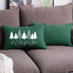 May include: A rectangular green throw pillow featuring a white graphic print of seven stylized evergreen trees of varying designs. Below the trees, the white cursive text reads "oh Christmas Tree". The pillow has a textured fabric finish.