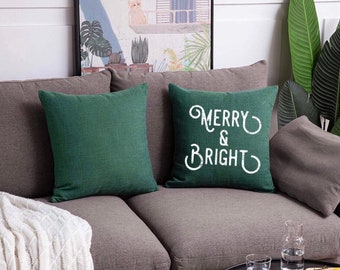 Puede incluir: Dos cojines cuadrados con una funda de tela verde oscuro texturizada. Un cojín es liso, mientras que el otro presenta el texto impreso en blanco "MERRY & BRIGHT" en una fuente decorativa estilizada centrada en la parte frontal del cojín.