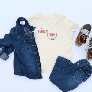 K&ouml;nnte beinhalten: Eine Zusammenstellung von Kinderkleidung, darunter ein Jeanshemd, ein cremefarbenes T-Shirt mit dem Text "HE HAS RISEN/JUST AS HE SAID", eine dunkelblaue Jeans und grau-braune Schuhe. Die Artikel sind auf wei&szlig;em Grund angeordnet.
