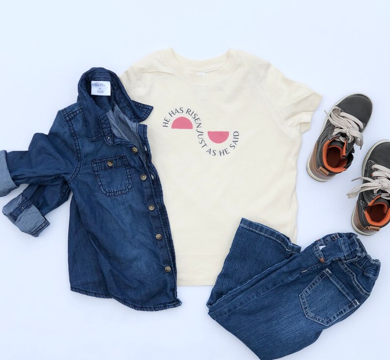K&ouml;nnte beinhalten: Eine Zusammenstellung von Kinderkleidung, darunter ein Jeanshemd, ein cremefarbenes T-Shirt mit dem Text "HE HAS RISEN/JUST AS HE SAID", eine dunkelblaue Jeans und grau-braune Schuhe. Die Artikel sind auf wei&szlig;em Grund angeordnet.