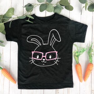 May include: Black toddler t-shirt with a white bunny face wearing pink glasses. The shirt is decorated with a bunny face graphic. Two orange carrots and green foliage are placed around the shirt on a white wooden surface.