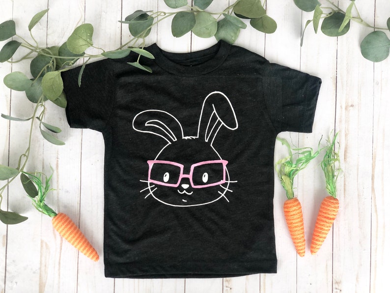 May include: Black toddler t-shirt with a white bunny face wearing pink glasses. The shirt is decorated with a bunny face graphic. Two orange carrots and green foliage are placed around the shirt on a white wooden surface.
