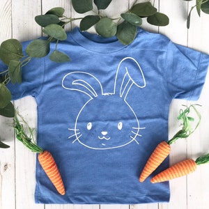 May include: A blue t-shirt with a white bunny face design. The shirt is surrounded by green leaves and three orange and green carrot decorations. The shirt is a children's apparel item.