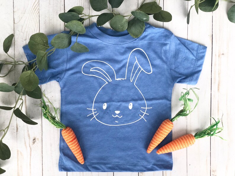 May include: A blue t-shirt with a white bunny face design. The shirt is surrounded by green leaves and three orange and green carrot decorations. The shirt is a children's apparel item.