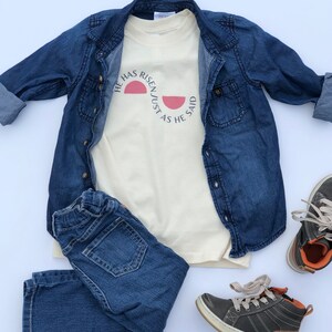 K&ouml;nnte beinhalten: Ein cremefarbenes T-Shirt mit dem Text "HE HAS RISEN/JUST AS HE SAID" in einem rosa Halbkreis-Design. Das Shirt wird mit einer blauen Jeansjacke, einer blauen Jeans und grauen und braunen Turnschuhen kombiniert, was ein l&auml;ssiges Outfit ergibt.