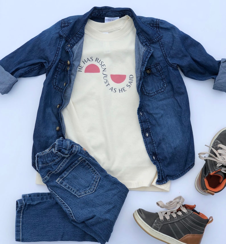 K&ouml;nnte beinhalten: Ein cremefarbenes T-Shirt mit dem Text "HE HAS RISEN/JUST AS HE SAID" in einem rosa Halbkreis-Design. Das Shirt wird mit einer blauen Jeansjacke, einer blauen Jeans und grauen und braunen Turnschuhen kombiniert, was ein l&auml;ssiges Outfit ergibt.