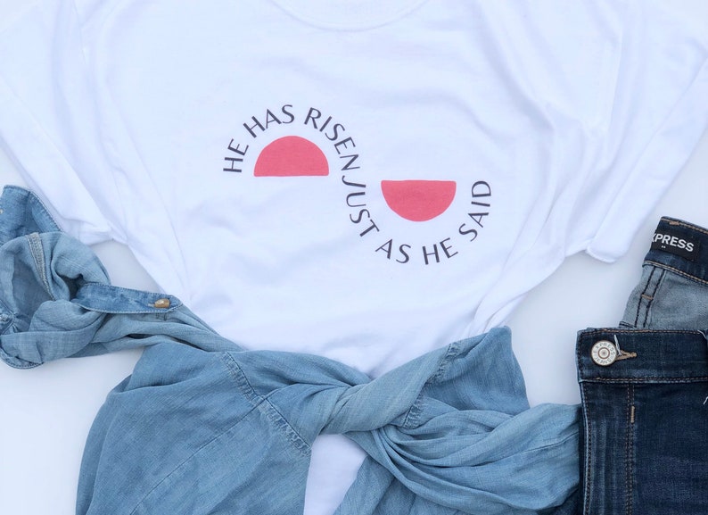 K&ouml;nnte beinhalten: Wei&szlig;es T-Shirt mit dem schwarzen Text "HE HAS RISEN JUST AS HE SAID" und einem roten Halbkreis-Design. Ein hellblaues Jeanshemd und Jeans sind ebenfalls im Bild.