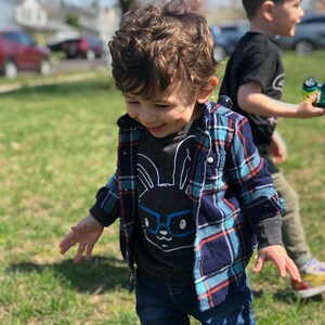 May include: A young child wearing a gray graphic tee with a bunny wearing glasses, a blue and navy plaid shirt, and blue jeans. The child is outside on a sunny day, with another child in the background.