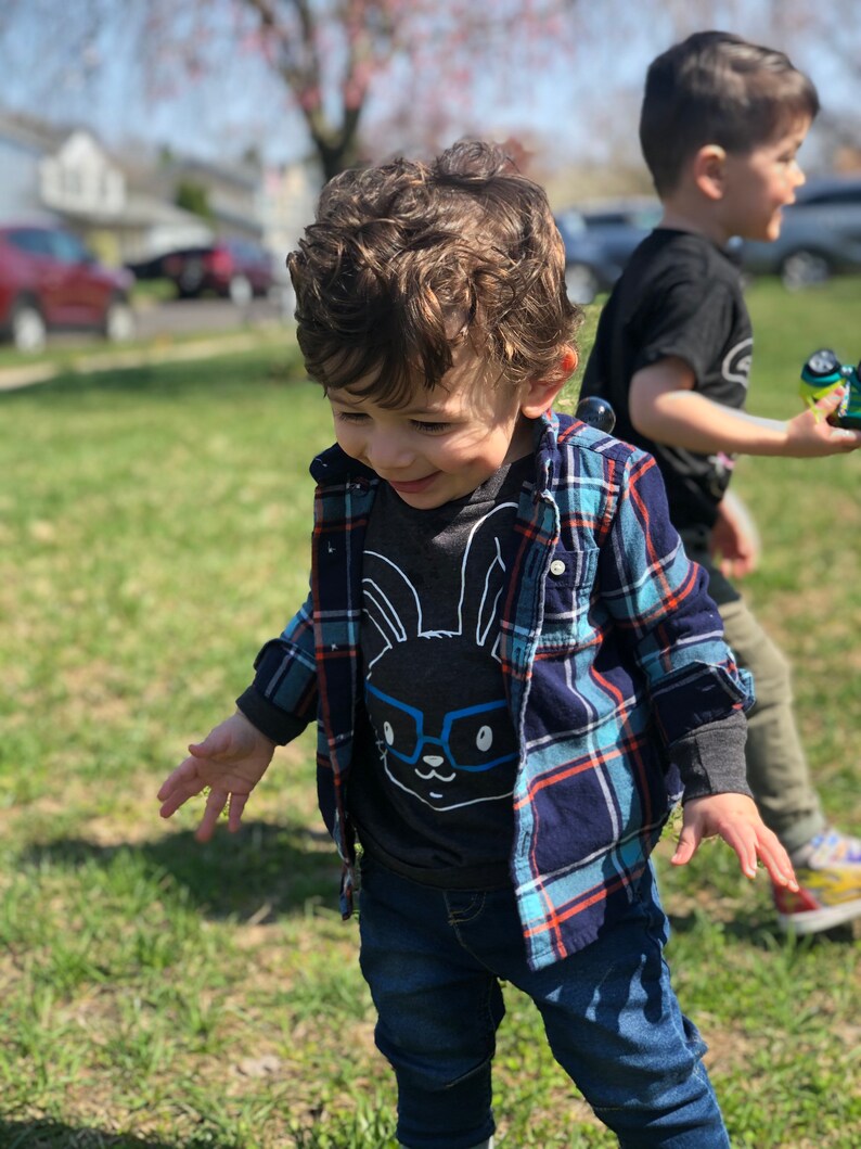 May include: A young child wearing a gray graphic tee with a bunny wearing glasses, a blue and navy plaid shirt, and blue jeans. The child is outside on a sunny day, with another child in the background.
