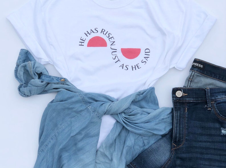 K&ouml;nnte beinhalten: Wei&szlig;es T-Shirt mit dem Text "HE HAS RISEN JUST AS HE SAID" in Schwarz, mit einer roten Sonnengrafik. Ein hellblaues Jeanshemd und eine dunkelblaue Jeans sind ebenfalls im Bild. Das Hemd ist gefaltet und mit der restlichen Kleidung gestylt.