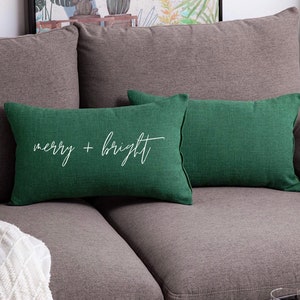 May include: Two rectangular throw pillows in a deep forest green color with a textured fabric finish. The front pillow features the white cursive text "merry + bright" centered on its surface. Both pillows have a simple, clean-edged design.