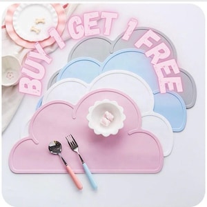 May include: Four silicone placemats shaped like clouds in pink, white, light blue, and gray. The placemats are stacked on top of each other. The top placemat is pink and has a small white bowl with two pink marshmallows in it. Two silver forks and a silver spoon are on the pink placemat. The image has the text "Buy 1 Get 1 Free" in pink letters.