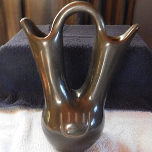 Może przedstawiać: Czarny ceramiczny wazon z dwoma wylewkami i uchwytem. Wazon ma gładkie, błyszczące wykończenie i ręcznie wykonany wzór na spodzie.