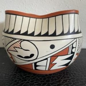 May include: A hand-painted ceramic bowl with a unique shape and intricate geometric designs. The bowl features a cream-colored background with black, red, and white patterns. The rim is a reddish-brown color.