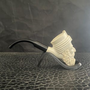 Puede incluir: Una pipa de tabaco color crema con un diseño de rostro tallado, un vástago negro y un soporte negro. La pipa presenta una representación detallada de un hombre barbudo con turbante. La pipa descansa sobre un soporte negro.