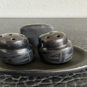 May include: A black ceramic salt and pepper set with a matching toothpick holder. The salt and pepper shakers have a rounded shape with decorative etched patterns. The set sits on a round tray.