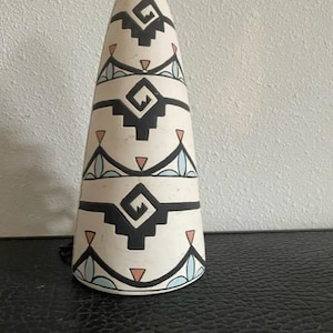 Peut inclure: Un objet décoratif conique en céramique blanc cassé, orné d'un motif d'inspiration amérindienne. Le motif présente des formes géométriques noires, des triangles et des lignes courbes bleues et roses. Il y a une petite boucle en haut.