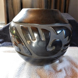 May include: Black pottery vase with a carved design. The vase has a rounded shape and a wide mouth.