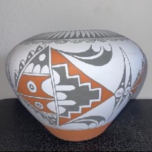 Puede incluir: Una olla de cerámica blanca con forma ancha y redondeada. Presenta diseños geométricos en naranja, gris y negro. La base es naranja y el borde tiene un patrón decorativo. La olla es probablemente hecha a mano y muestra artesanía tradicional.