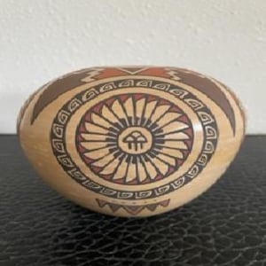 以下が含まれることがあります： 手描きの楕円形陶器ボウル。ベースは明るいベージュ色。ボウルの中央には、茶色と赤色の放射線状のラインを持つ円形デザインがあり、幾何学模様で囲まれています。縁には茶色のボーダーと装飾的な要素があります。