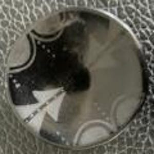 May include: A silver-toned, circular button with an etched design. The design features a series of curved lines, dots, and arrow-like shapes. The button has a slightly domed surface and a reflective quality.