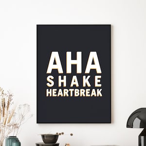 May include: A framed print with the words "AHA SHAKE HEARTBREAK" in white with a gold outline, set against a dark navy background. The print is above a wooden cabinet with decorative items.