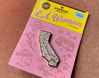 May include: Gold enamel pin shaped like the state of California with the text "LA Woman" inside. The pin is on a pink card with the text "Sunrise Motel" and "Eclectic Goods".