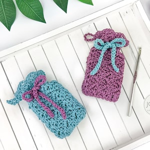 Puede incluir: Dos bolsas de crochet con cordón en verde azulado y morado, con lazos verde azulado y morados. Un ganchillo plateado y dos ovillos de lana también son visibles en una bandeja de madera blanca. Las bolsas están hechas a mano y tienen una puntada texturizada.