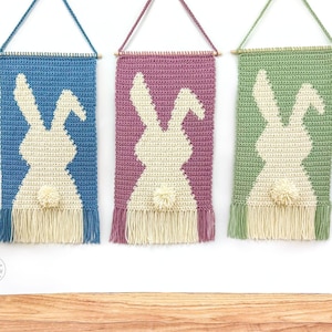 Könnte beinhalten: Drei gehäkelte Wandbehänge in Blau, Lila und Grün, jeweils mit einer weißen Hasen-Silhouette. Die Aufhänger haben am unteren Ende Fransen und werden von einem Holzstab und einer passenden Kordel gehalten. Ein Bommelschwanz sorgt für einen verspielten Touch.