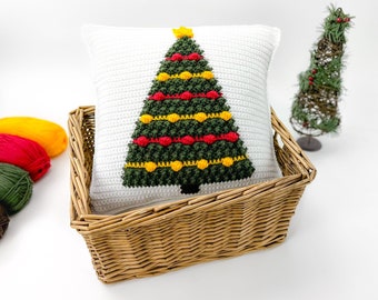 May include: A white crocheted pillow with a green Christmas tree design. The tree is decorated with red and yellow ornaments. The pillow is in a wicker basket.