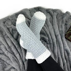 May include: A pair of cozy, light blue and white knit socks. The socks are resting on a soft, gray blanket. The socks have a textured pattern and white accents at the toes and cuffs. The blanket has a plush, furry texture.