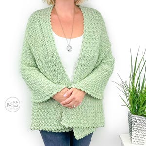 May include: A light sage green crochet cardigan featuring a textured stitch pattern, long sleeves, and an open front design. The garment is worn over a plain white top and includes a silver pendant necklace. A logo is visible on the lower left.
