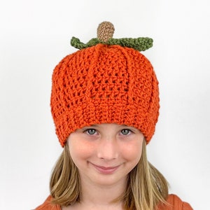 K&ouml;nnte beinhalten: Eine geh&auml;kelte K&uuml;rbis-M&uuml;tze in Orange mit einem gr&uuml;nen Stiel und Blatt. Die M&uuml;tze wird von einem Kind mit blonden Haaren getragen.