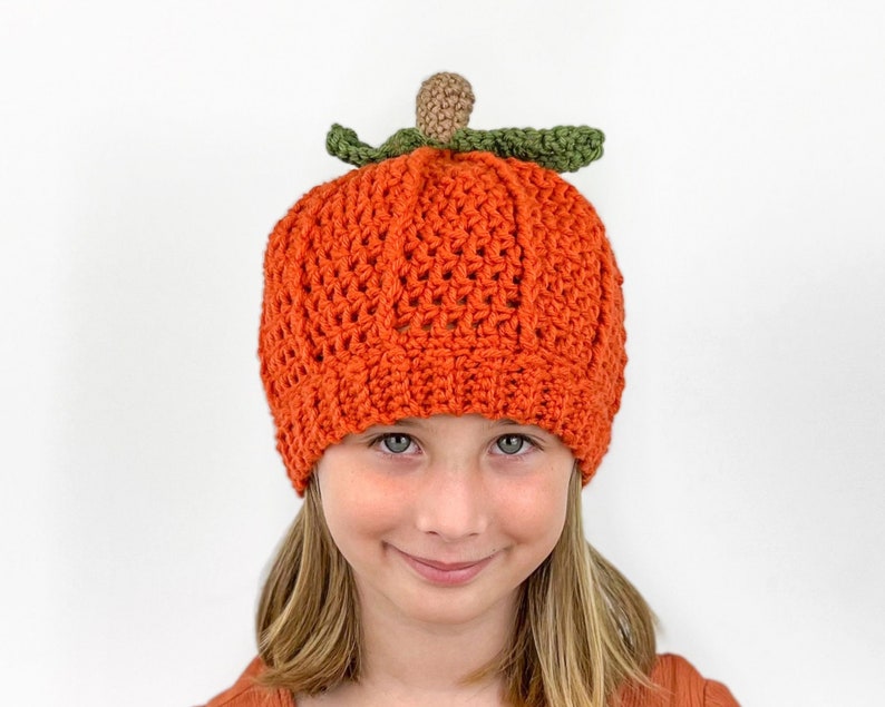 K&ouml;nnte beinhalten: Eine geh&auml;kelte K&uuml;rbis-M&uuml;tze in Orange mit einem gr&uuml;nen Stiel und Blatt. Die M&uuml;tze wird von einem Kind mit blonden Haaren getragen.