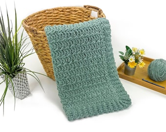 May include: A soft, seafoam green crochet blanket with a textured stitch pattern. The blanket is displayed near a woven basket, a small bouquet of flowers, and a ball of yarn. The blanket's dimensions are not visible.