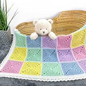 Puede incluir: Una manta de bebé de ganchillo con un borde blanco y un esquema de color pastel. La manta está hecha de cuadrados en tonos de rosa, amarillo, verde y azul. Un osito de peluche está sentado encima de la manta.