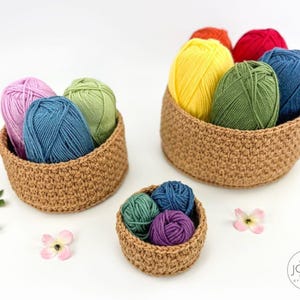 May include: Three crocheted yarn baskets filled with colorful yarn balls. The yarn colors include pink, blue, green, yellow, orange, and red. The baskets are a light brown color and are arranged on a white surface.